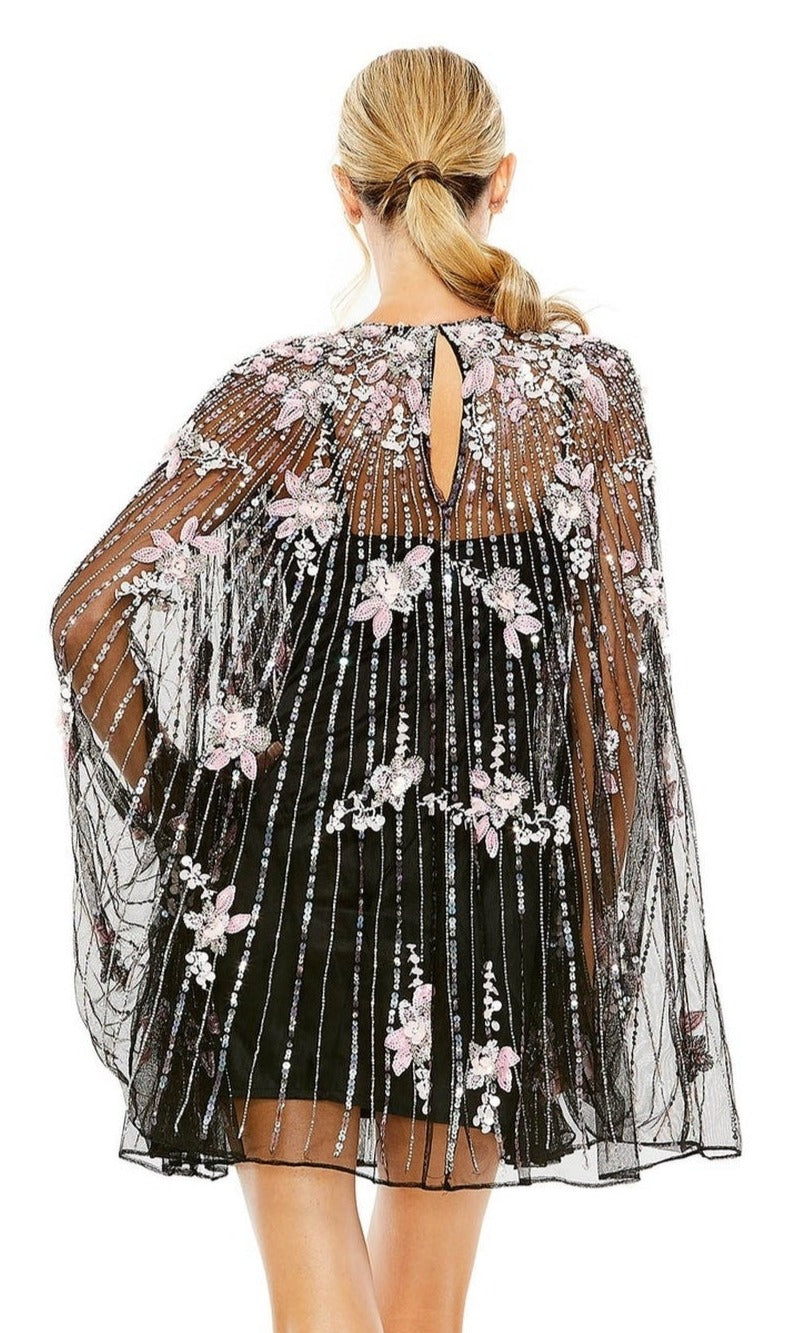 A woman with blonde hair in a low ponytail is seen from the back wearing the Mac Duggal 5917 Beaded-Cape Short Hoco Dress, featuring a black dress with a sheer, floral-embroidered cape adorned with pink and white flowers.